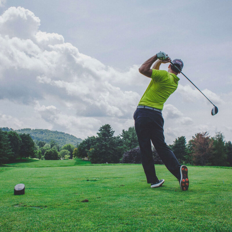 A golfer in a yellow shirt and dark pants swings a golf club on a lush green course surrounded by trees, with hills and a partly cloudy sky.