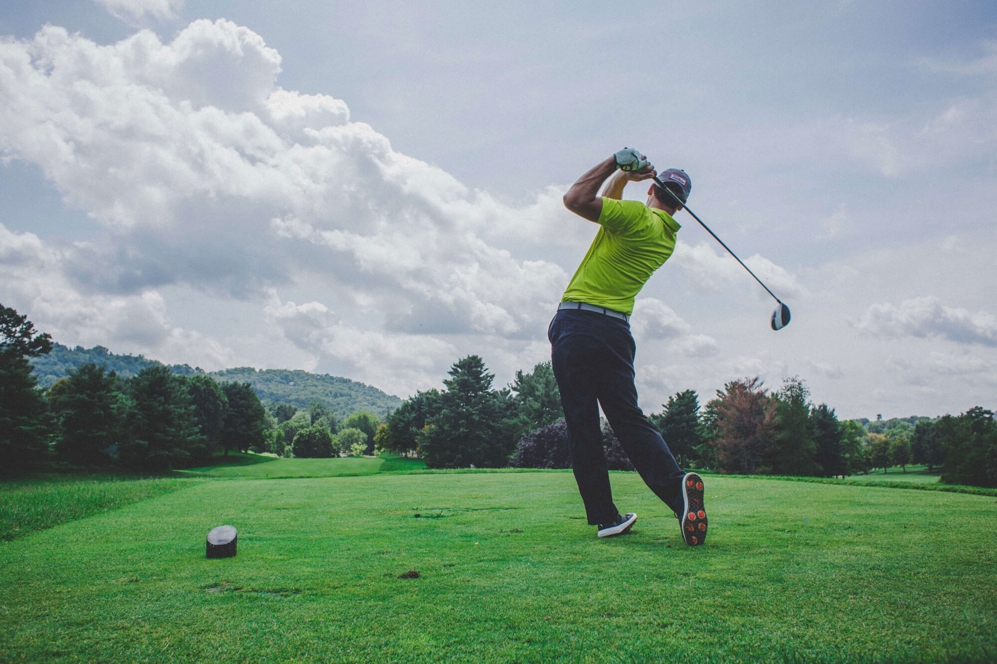 A golfer in a yellow shirt and dark pants swings a golf club on a lush green course surrounded by trees, with hills and a partly cloudy sky.