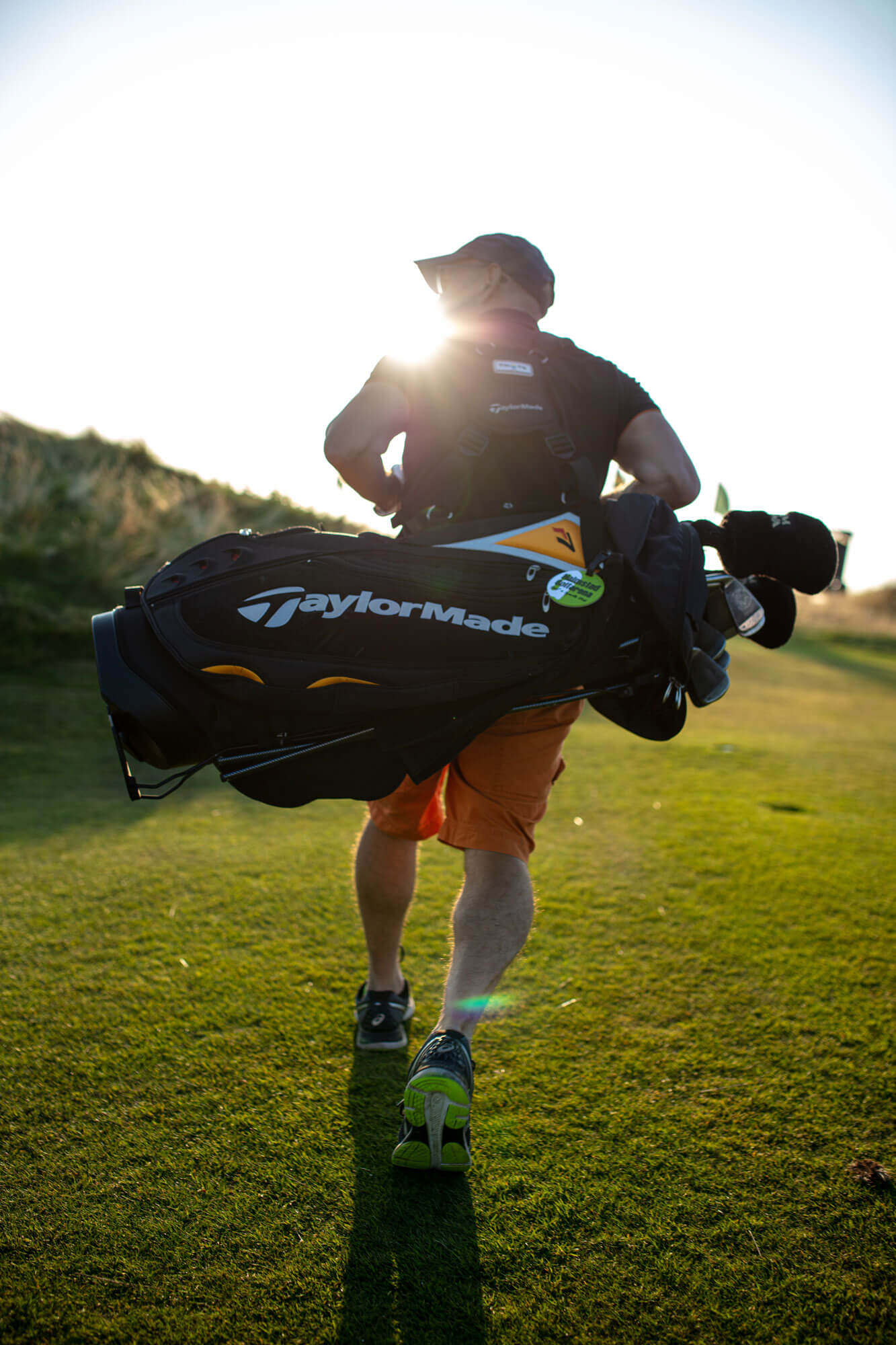 A person walks on a sunny golf course in London carrying a large TaylorMade golf bag over their shoulder, the sun shining brightly.