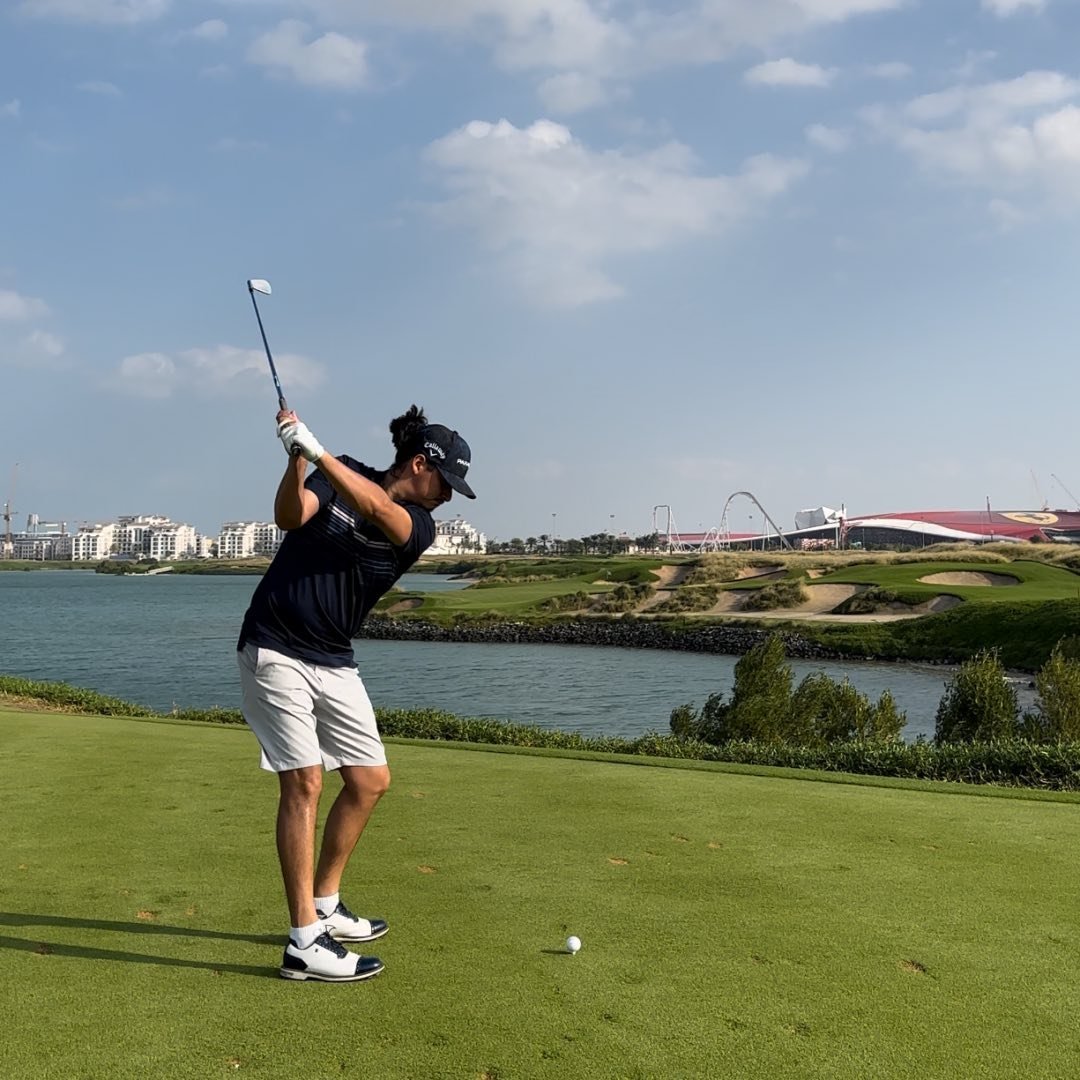A golfer in a navy shirt and white shorts prepares to swing on a green golf course near a river in London, with buildings, a roller coaster, and a partly cloudy sky in the background—perfect for those seeking golf memberships.