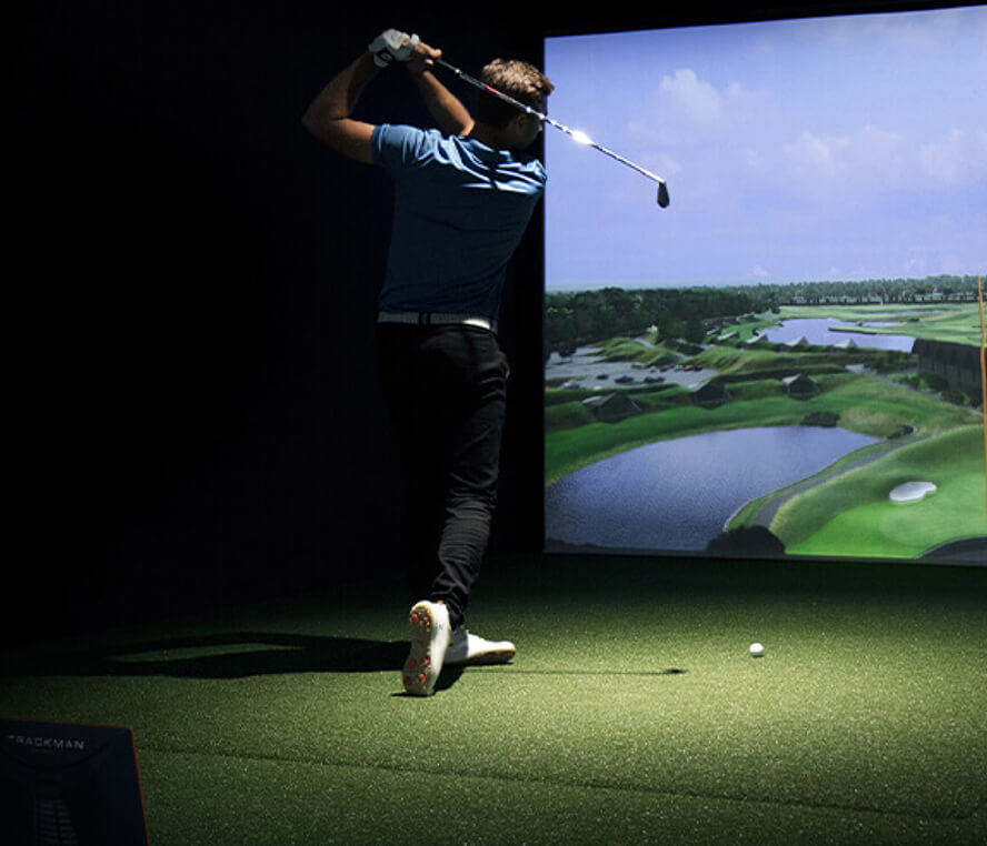 A golfer in mid-swing practices indoors on artificial turf, aiming at a large golf simulator screen displaying a virtual golf course—perfect for those seeking golf memberships or authentic golf London experiences year-round.