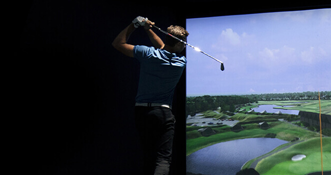 A person swings a golf club indoors in front of a large virtual screen displaying a realistic golf course with water and greenery.