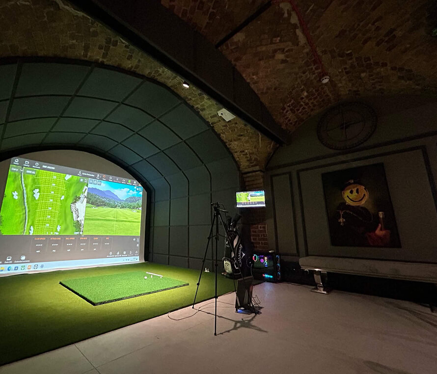 A golf simulator room with a large screen displaying a virtual golf course, a patch of artificial turf, and camera setup—perfect for private golf lessons. The room features brick arch ceilings and smiley face artwork on the wall.