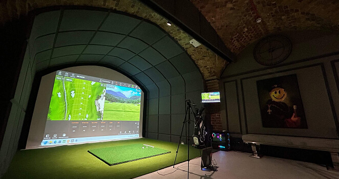 A golf simulator setup in a dimly lit room with brick arched ceiling offers virtual golf lessons on a large screen, green turf flooring, and computer equipment nearby. A painting and a clock hang on the wall.
