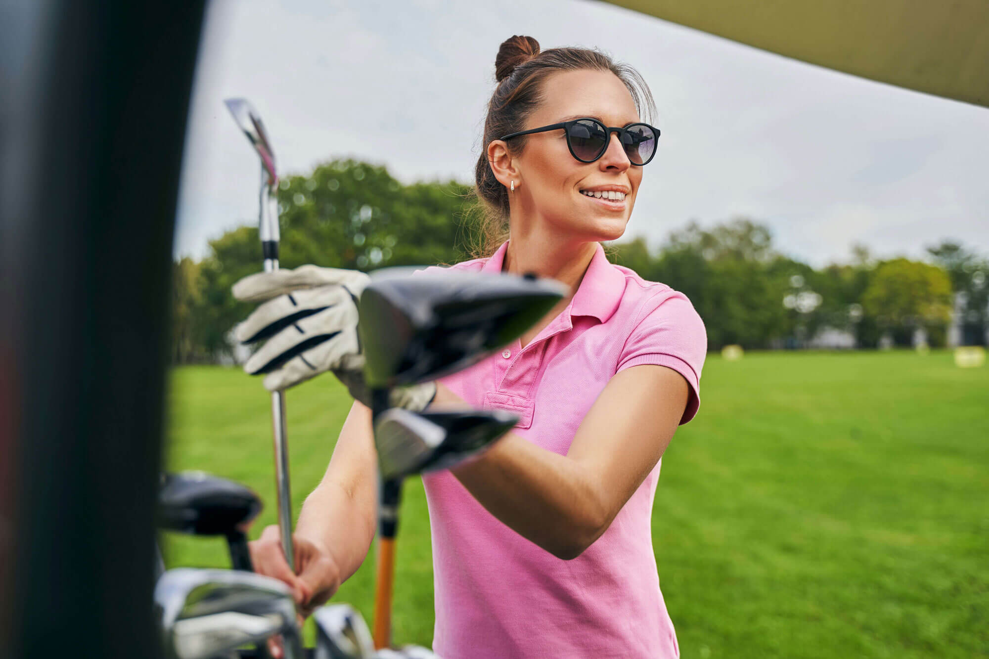 A woman wearing sunglasses and a pink polo shirt smiles while selecting a golf club from her golf bag on a green course, enjoying her golf lessons in London.