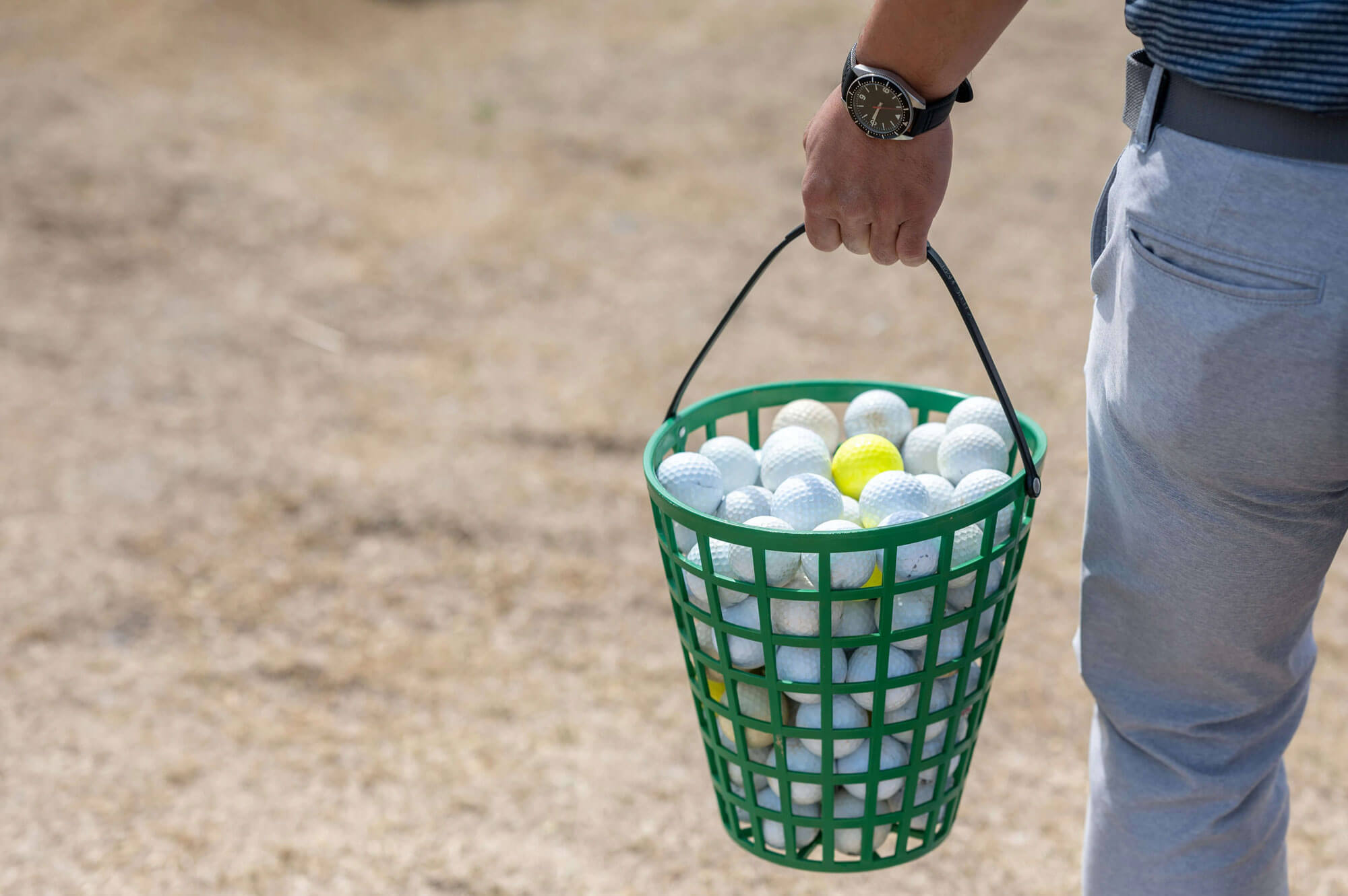 A person holding a green basket filled with white and yellow golf balls, standing on patchy grass—perfect for those considering golf London experiences or looking into golf lessons to improve their game. Only the persons torso and arm are visible.