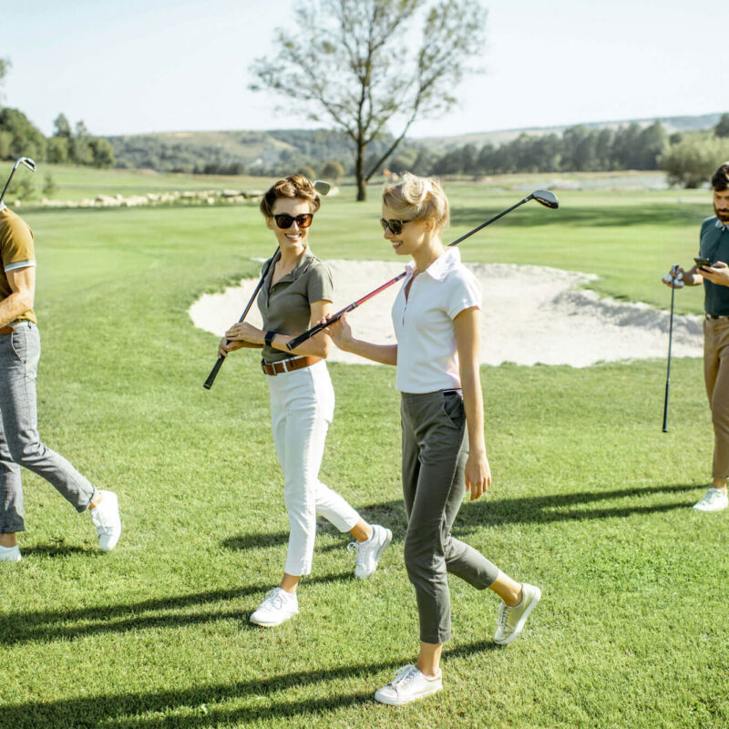 Four people enjoy a sunny day on a golf course in London—two women chatting with golf clubs, flanked by two men, one checking his mobile. Trees line the backdrop, perfectly capturing the spirit of golf in London and exclusive golf memberships.