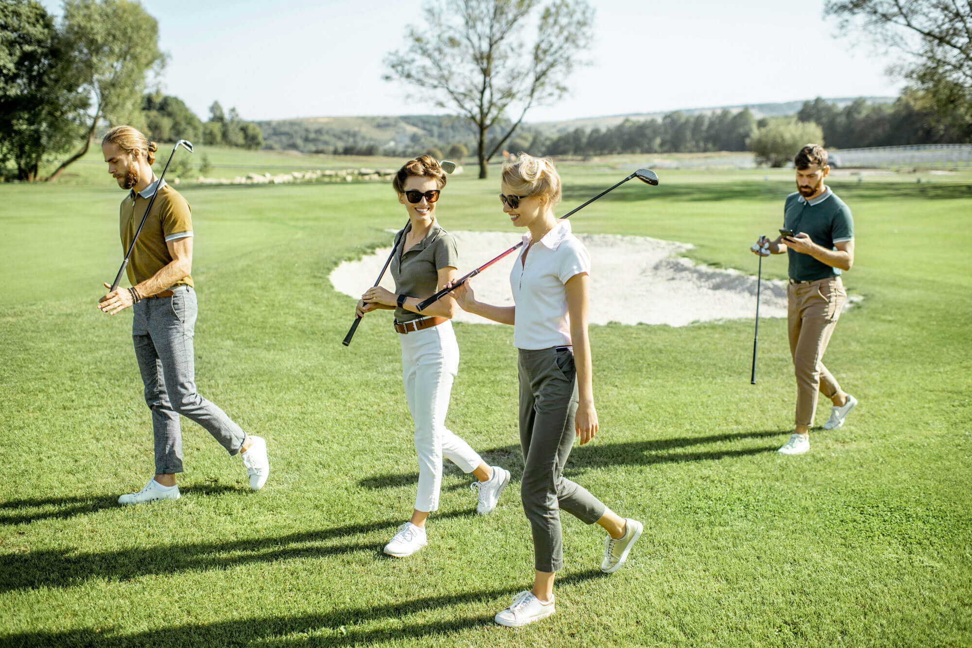Four people enjoy a sunny day on a golf course in London—two women chatting with golf clubs, flanked by two men, one checking his mobile. Trees line the backdrop, perfectly capturing the spirit of golf in London and exclusive golf memberships.