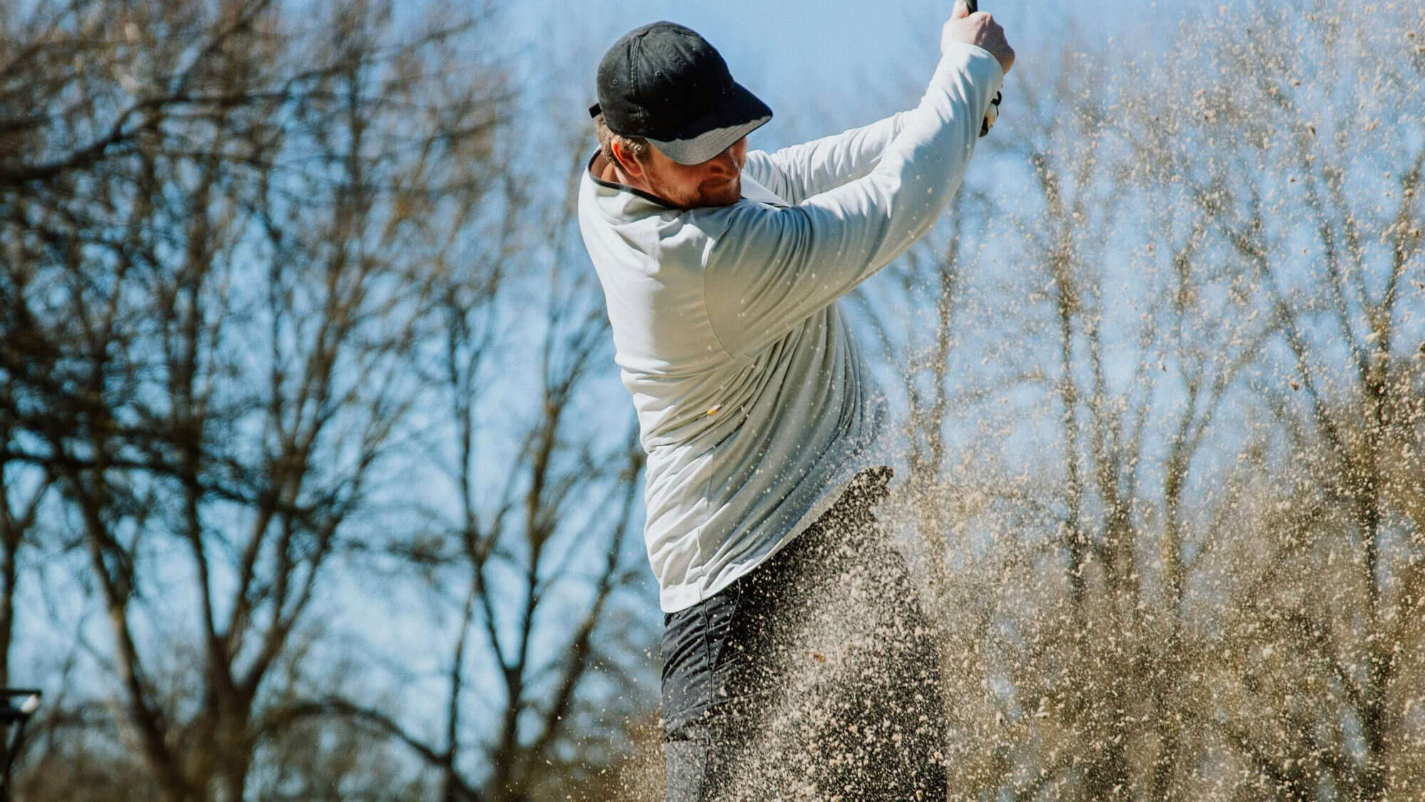 A person wearing a white long-sleeve shirt and black cap swings a golf club, sending sand flying—a perfect scene for those seeking golf lessons or golf memberships in scenic settings near golf London.