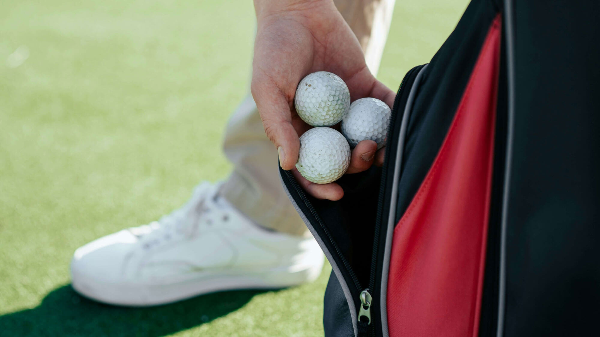A person holds four golf balls in one hand while taking them out of a red and black bag, with part of a white sneaker visible on the green—capturing the essence of golf on the course.