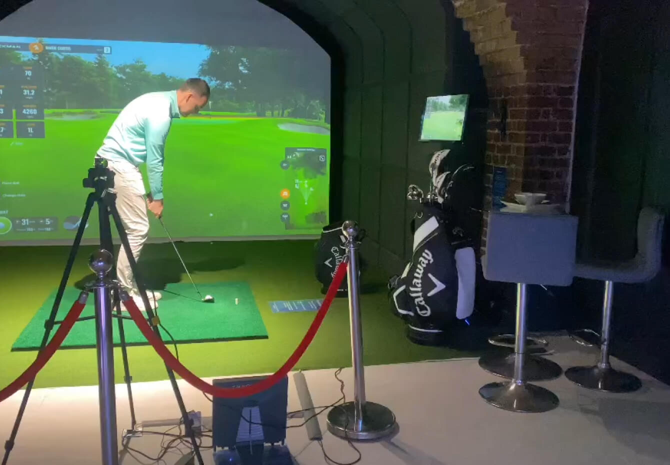 A man prepares to swing a golf club at an indoor golf simulator in London. Golf bags, clubs, and chairs sit nearby, while a red rope barrier frames the setup. The large screen displays a virtual golf course—perfect for golf lessons year-round.