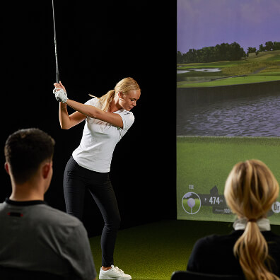 A woman swings a golf club in front of a golf simulator screen at Golf London, while two people sitting nearby watch her practice. The screen displays a virtual golf course, offering an immersive way to enjoy the game and explore golf memberships.