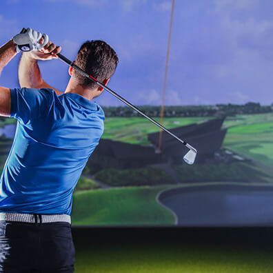 A person wearing a blue shirt swings a golf club indoors in London, facing a large screen displaying a virtual golf course with green fairways, water, and a bright sky—perfect for those interested in golf lessons or memberships.