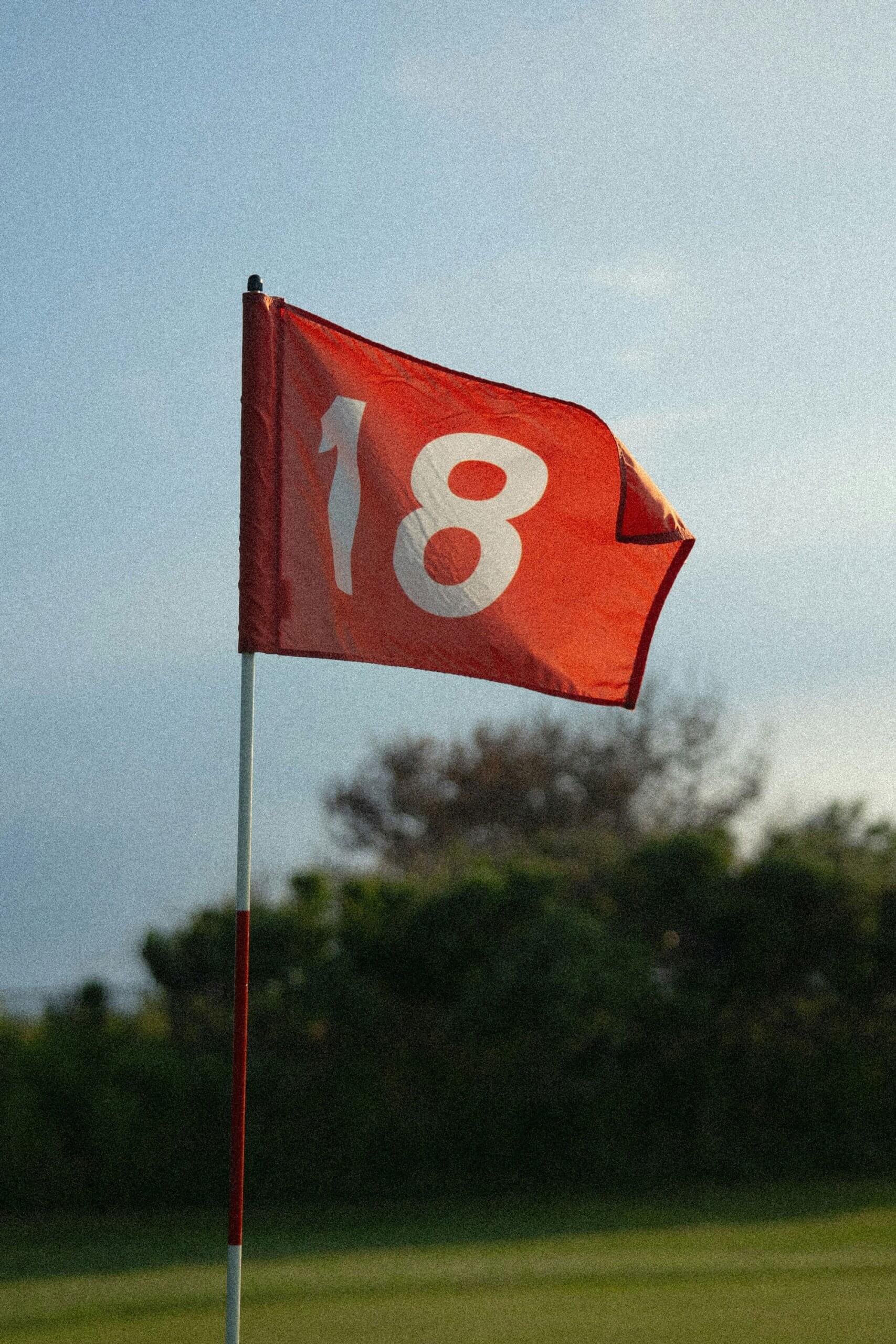 A red flag with the number 18 printed in white flies on a pole, marking the final hole of a golf course in London—an inviting scene for memorable golf lessons amid green grass and trees in the background.