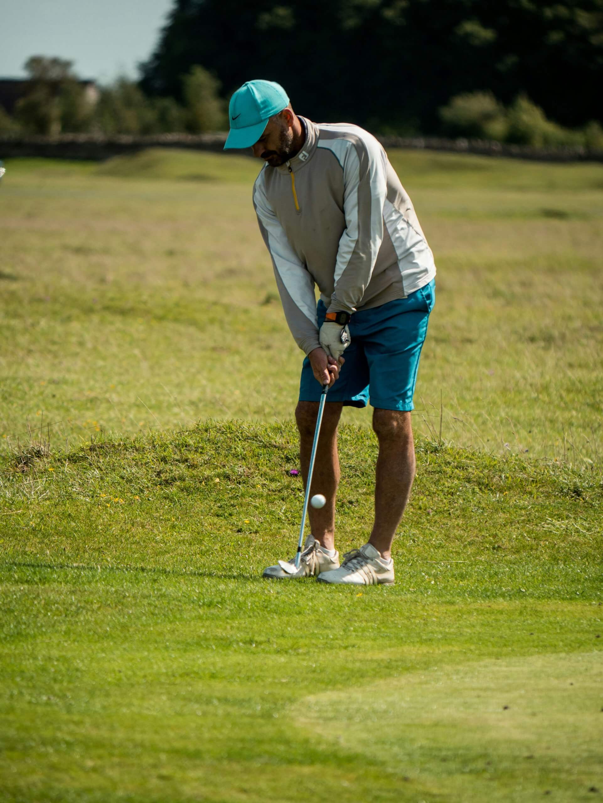 A man wearing a blue cap, gray jacket, and blue shorts is playing golf on a grassy course in London, focusing as he swings his club to hit the golf ball.