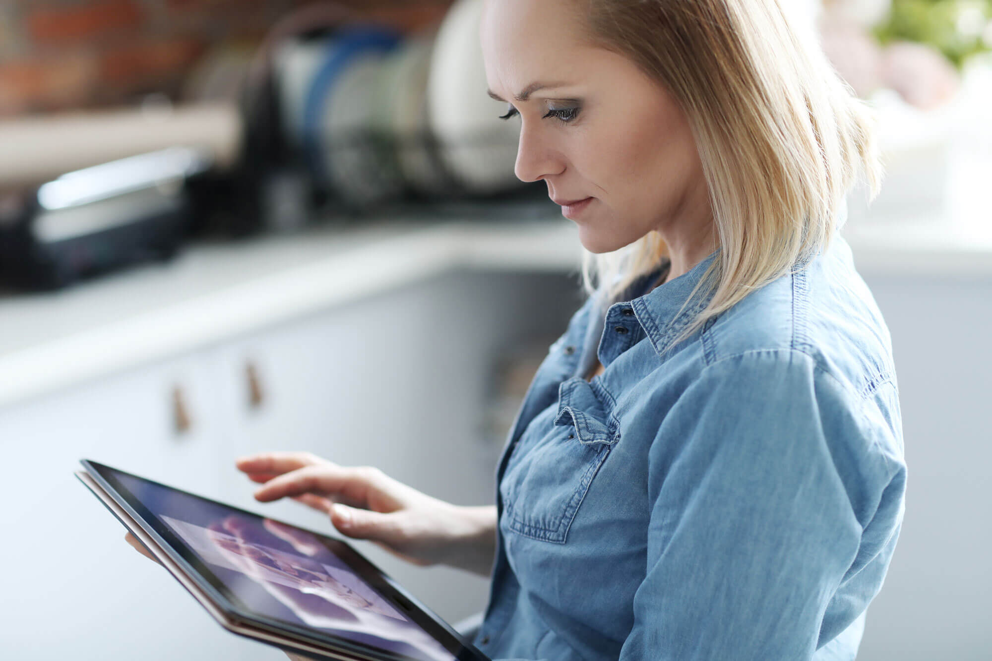 A woman in a denim shirt sits indoors, focused on her tablet as she explores golf memberships. The softly blurred background highlights her concentration.