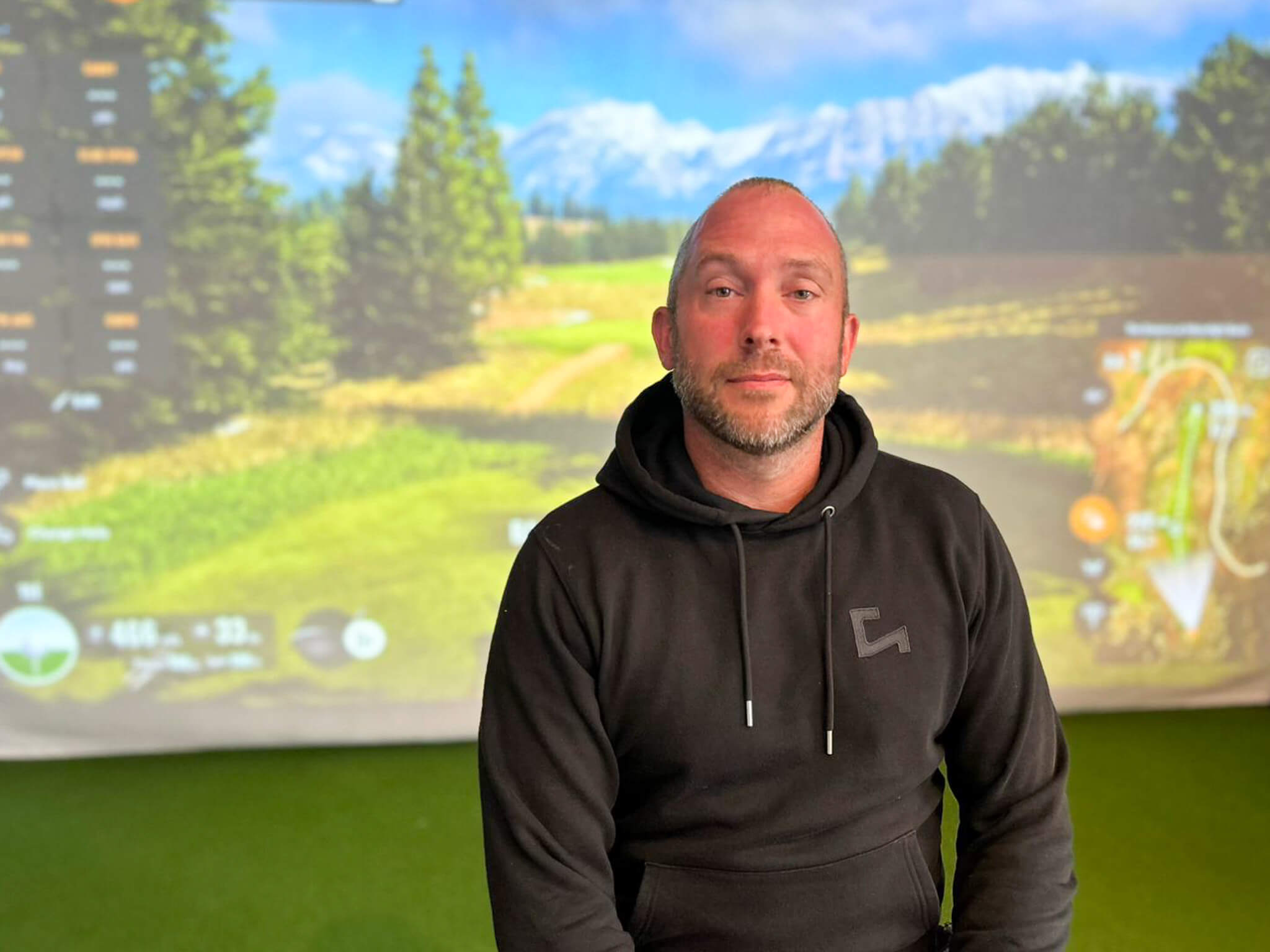 A man with a shaved head and trimmed beard wearing a black hoodie sits indoors in front of a TrackMan golf simulator screen displaying a scenic golf course, perfect for honing skills or enjoying his golf membership.