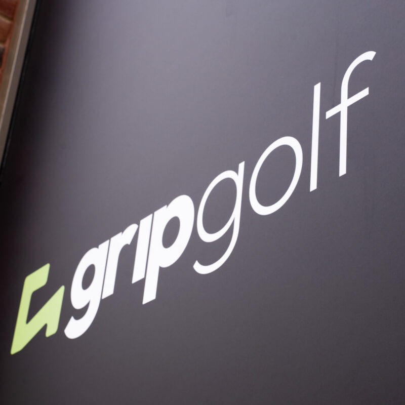 A black sign with the white text “gripgolf” and a green geometric logo to the left, mounted on a brick wall, signals a modern spot for golf lessons in London.