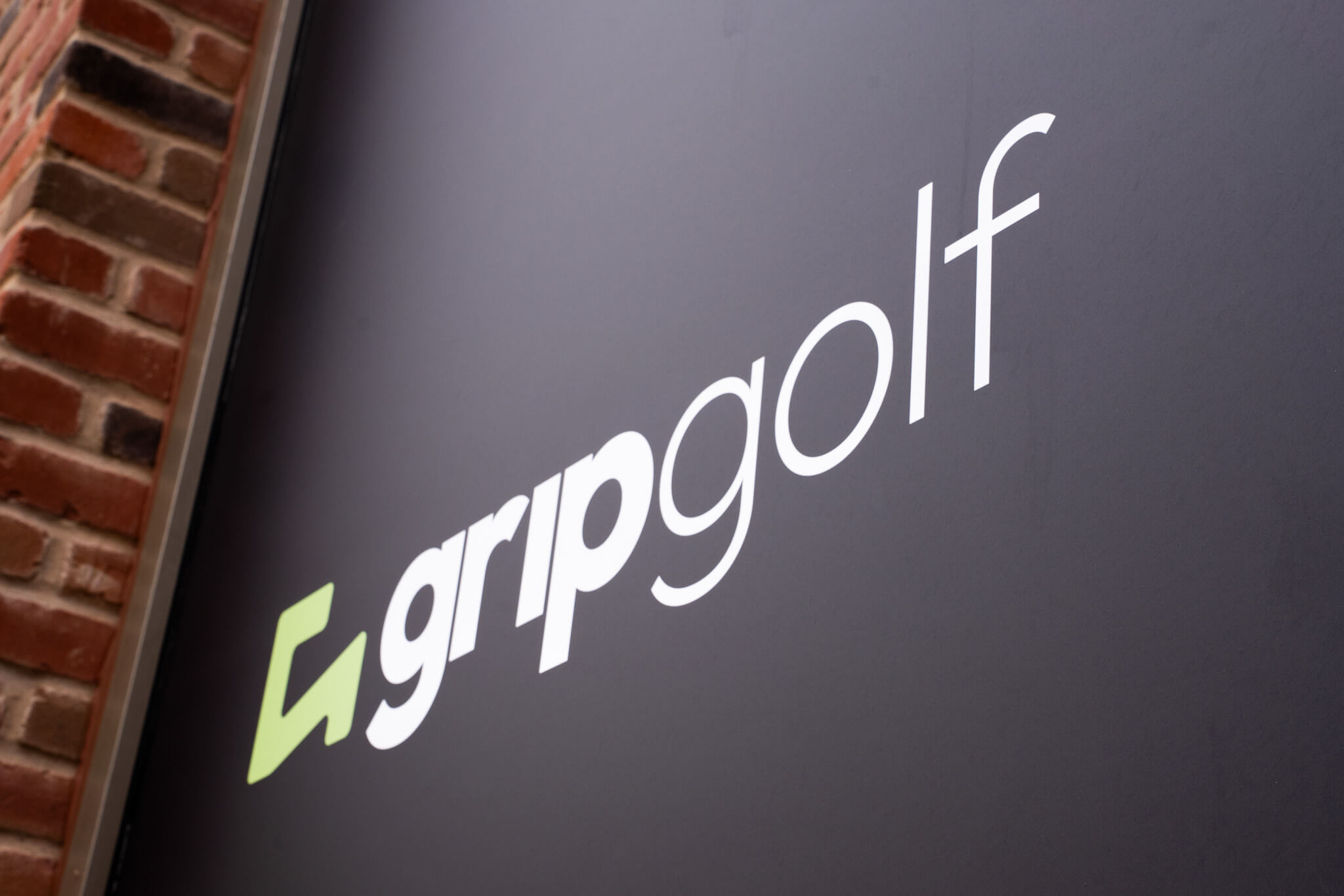 A black sign with the white text “gripgolf” and a green geometric logo to the left, mounted on a brick wall, signals a modern spot for golf lessons in London.