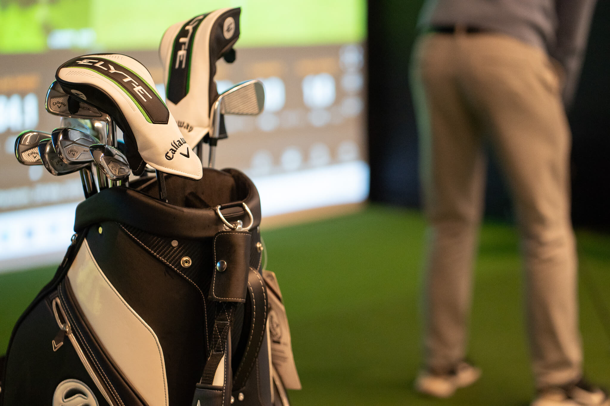 A close-up of a golf bag filled with clubs in the foreground, while a person in gray pants prepares to swing at a golf simulator screen—a perfect setting for golf lessons in London.