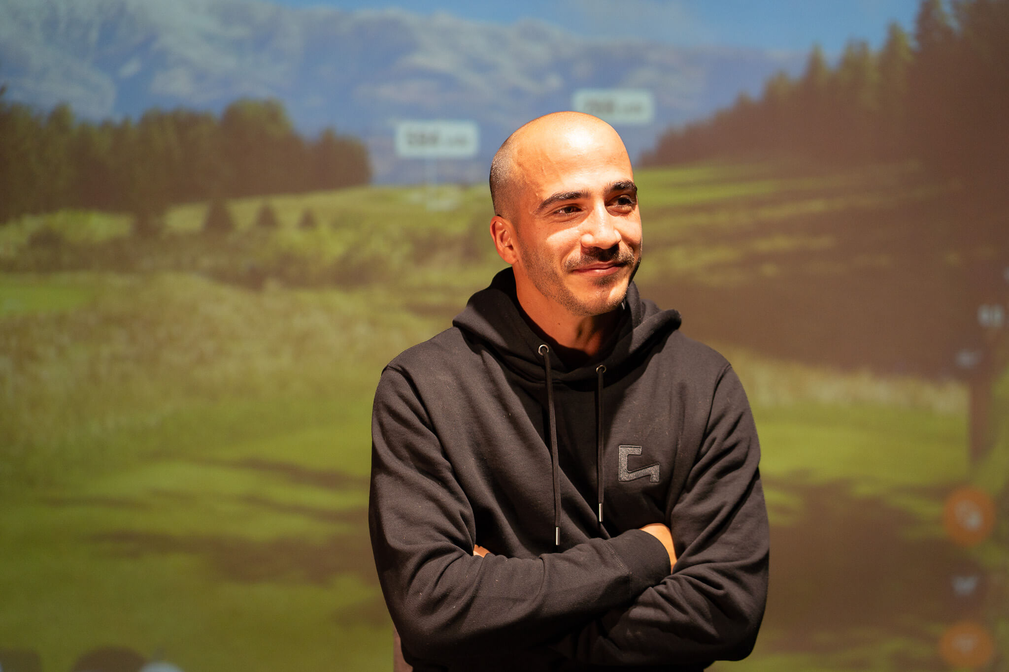 A man with a shaved head and mustache, wearing a dark hoodie, stands with his arms crossed and smiles in front of a blurred scenic background featuring green fields and trees—perfect for golf lessons or exploring golf membership options in London.