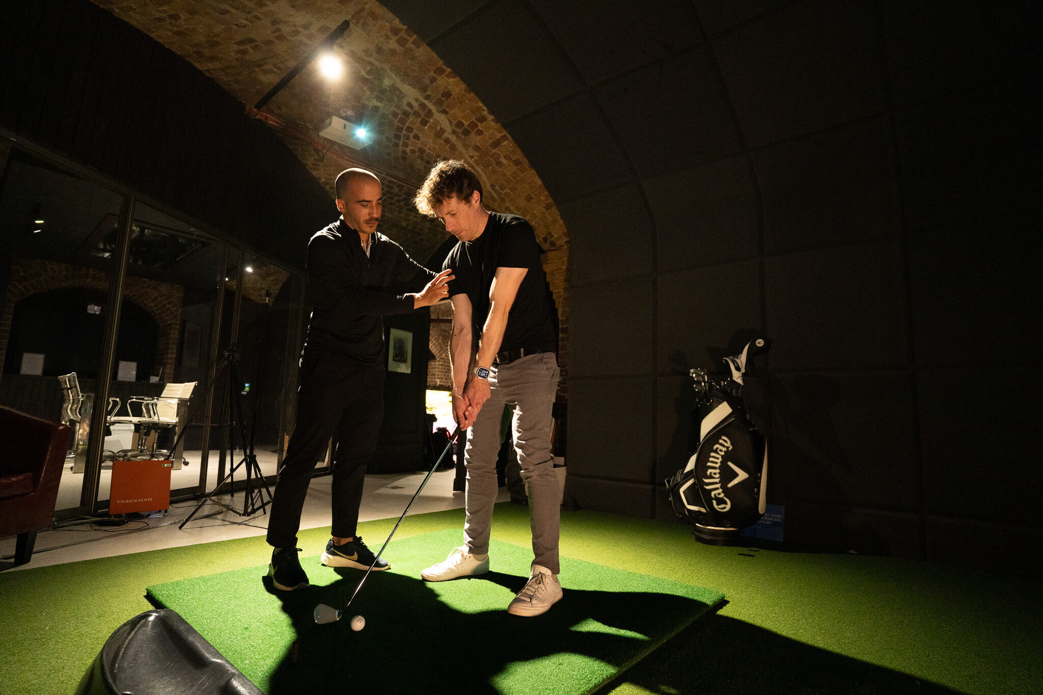 A golf instructor guides a student’s posture as he prepares to swing at an indoor golf facility in London, with artificial turf and golf equipment visible in the background. Consider a golf membership for expert coaching like this.