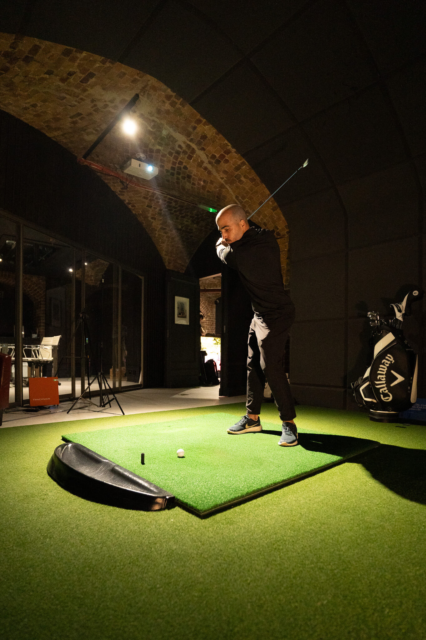 A man in dark clothing prepares to swing a golf club indoors on an artificial green mat, using Trackman technology. Golf equipment and brick archways enhance the stylish golf London atmosphere in the background.