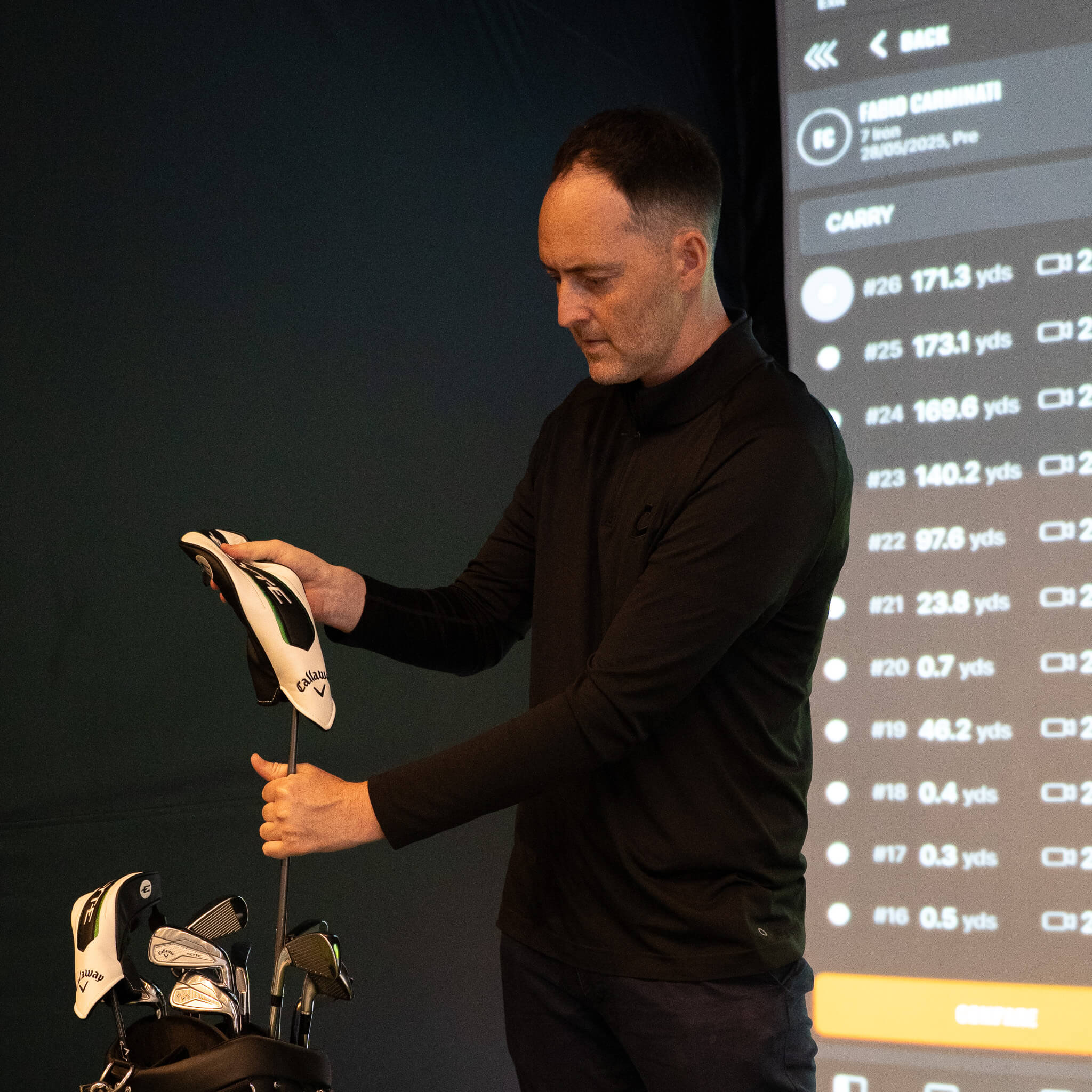 A man in a black shirt stands indoors, holding a golf club and placing a headcover on it. A bag of clubs is in front of him, and a digital screen displaying golf statistics hints at an advanced golf lesson—perhaps even in the heart of golf London.
