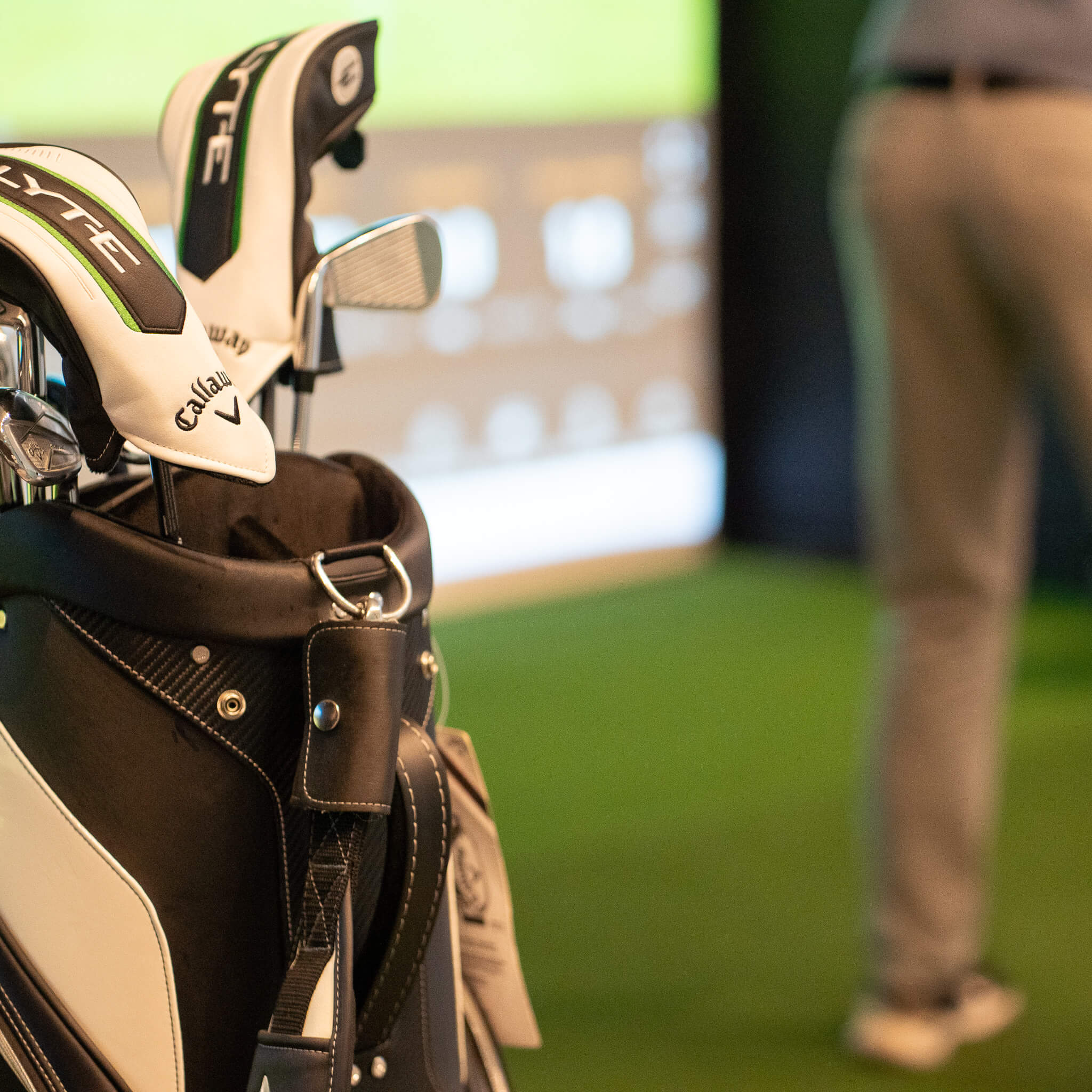 A close-up of a black and white golf bag holding Callaway clubs, with a person in light gray pants standing on green artificial turf at a golf London venue, and a blurred Trackman golf simulator screen in the background.