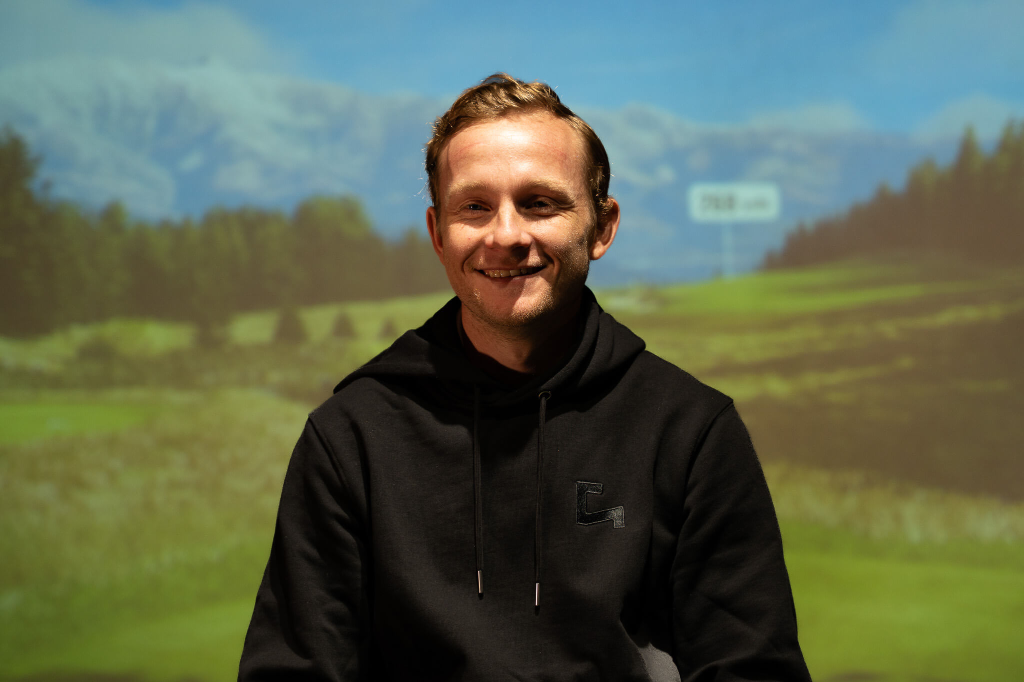 A young man with short light brown hair, wearing a black hoodie, smiles while sitting in front of a scenic landscape featuring green fields, trees, and mountains under a blue sky—perfect for a day of golf.