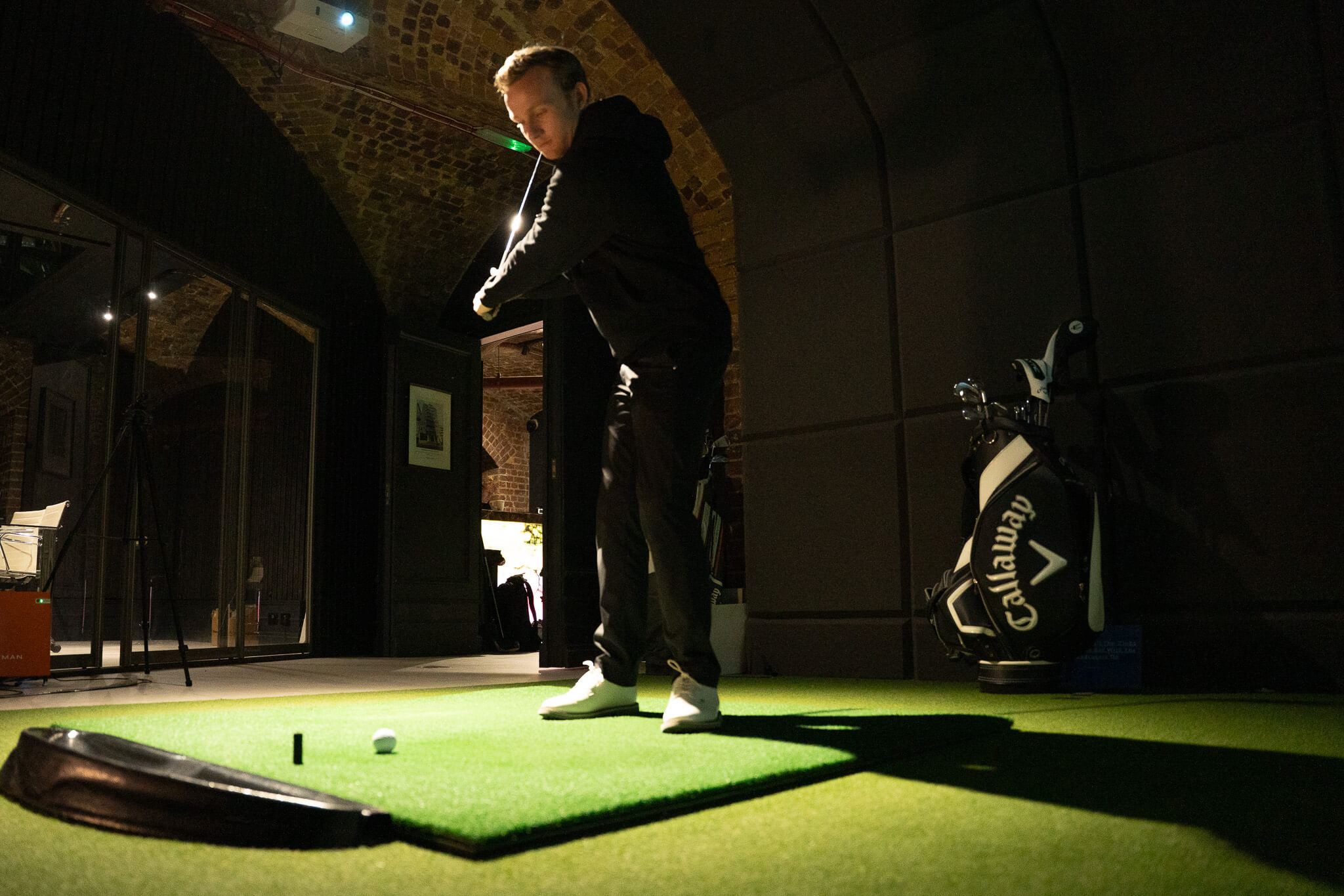 A person in dark clothing practices a golf swing indoors on a green mat, aiming at a golf ball. Golf clubs are seen in a Callaway bag nearby, capturing the essence of stylish indoor golf London sessions amid brick arches and dim lighting.