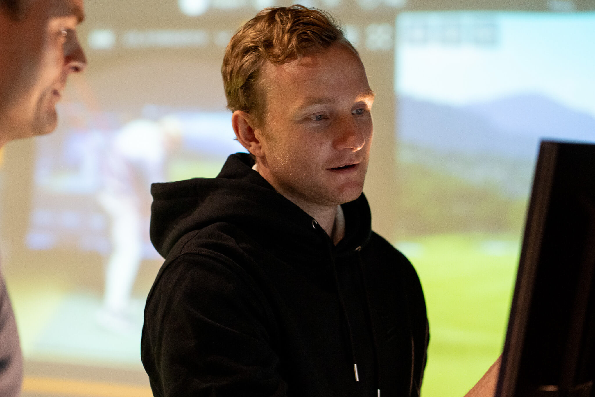 A man in a black hoodie looks at a computer screen, with another person partially visible beside him. The blurred background reveals a projected image of a person and landscape—hinting at indoor golf London practice with Trackman technology.