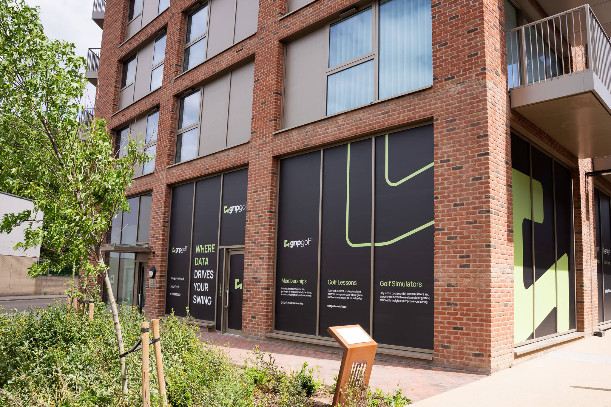 A brick building with large windows features black and green signage for LegaGolf, advertising golf memberships, golf lessons, and state-of-the-art golf simulators. Trees and a small planter are visible in the foreground.