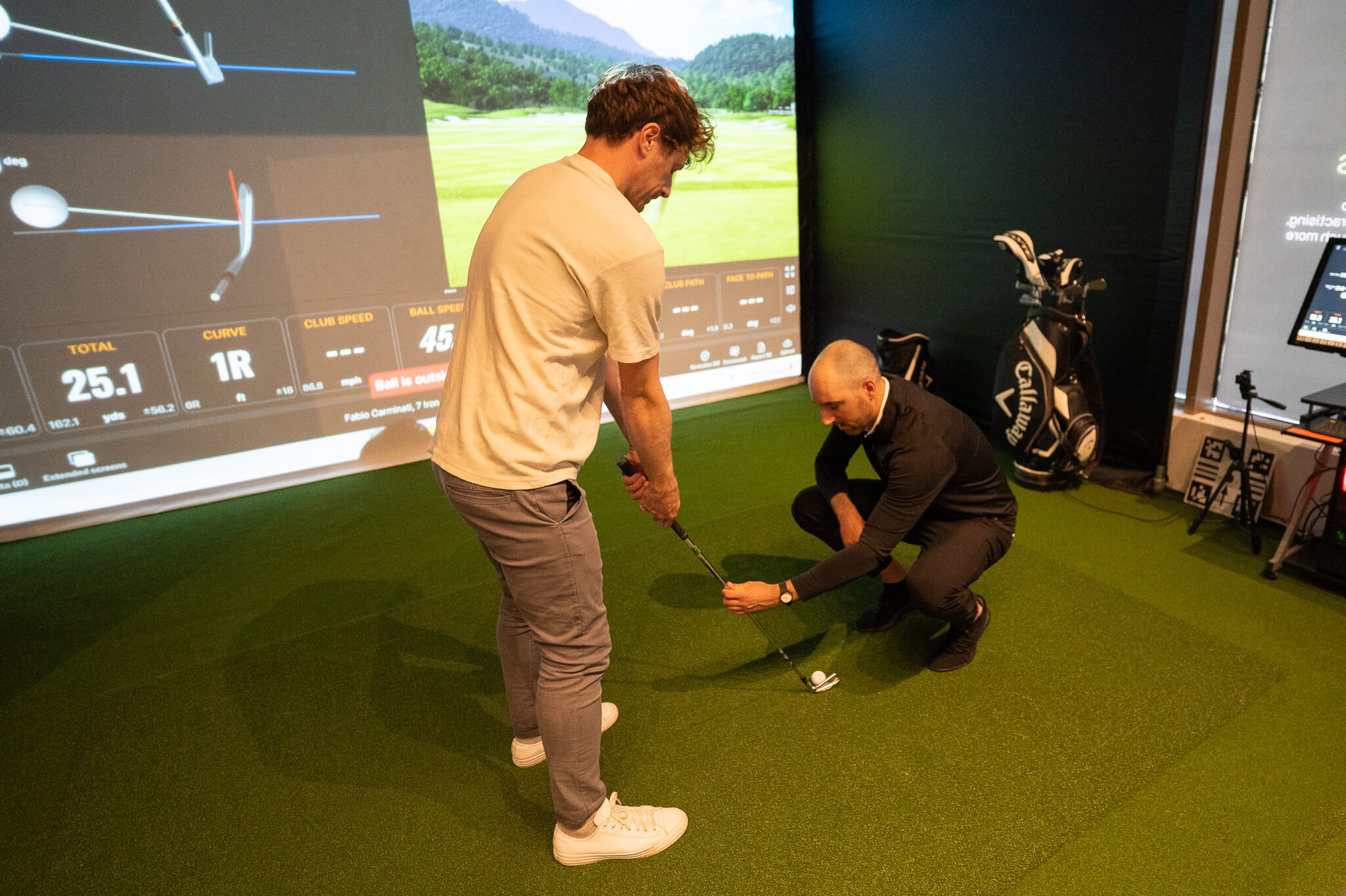 A golf instructor kneels on indoor turf, adjusting a student’s putter during golf lessons as the student prepares to swing. A TrackMan golf simulator screen and several golf clubs are visible in the background.