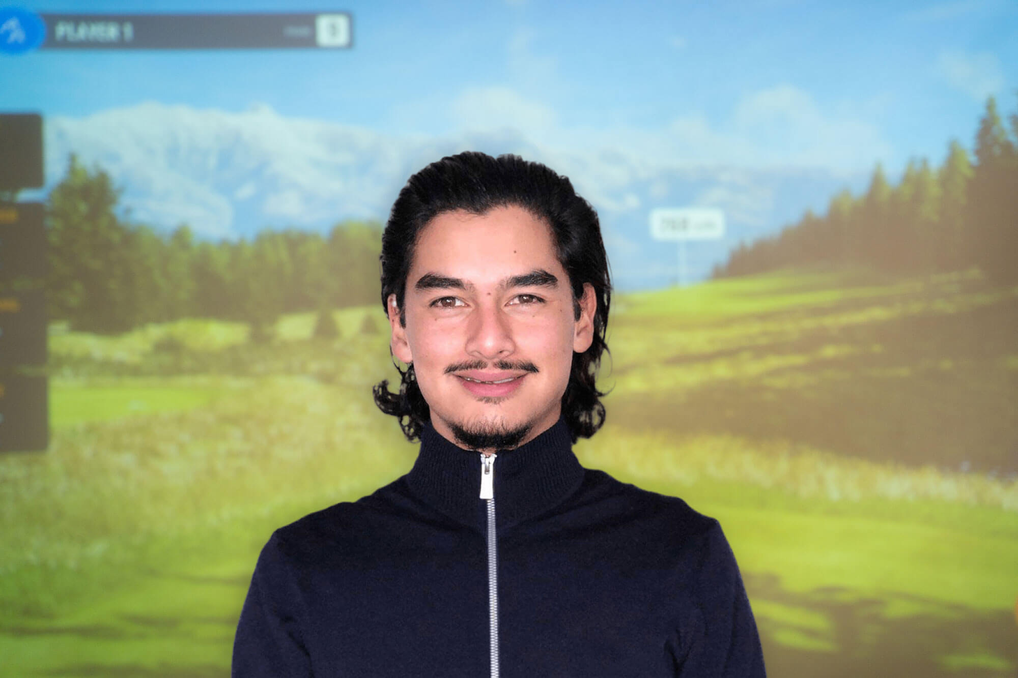 A man with dark hair and a trimmed beard, wearing a dark zip-up sweater, stands smiling in front of a blurred golf course background.