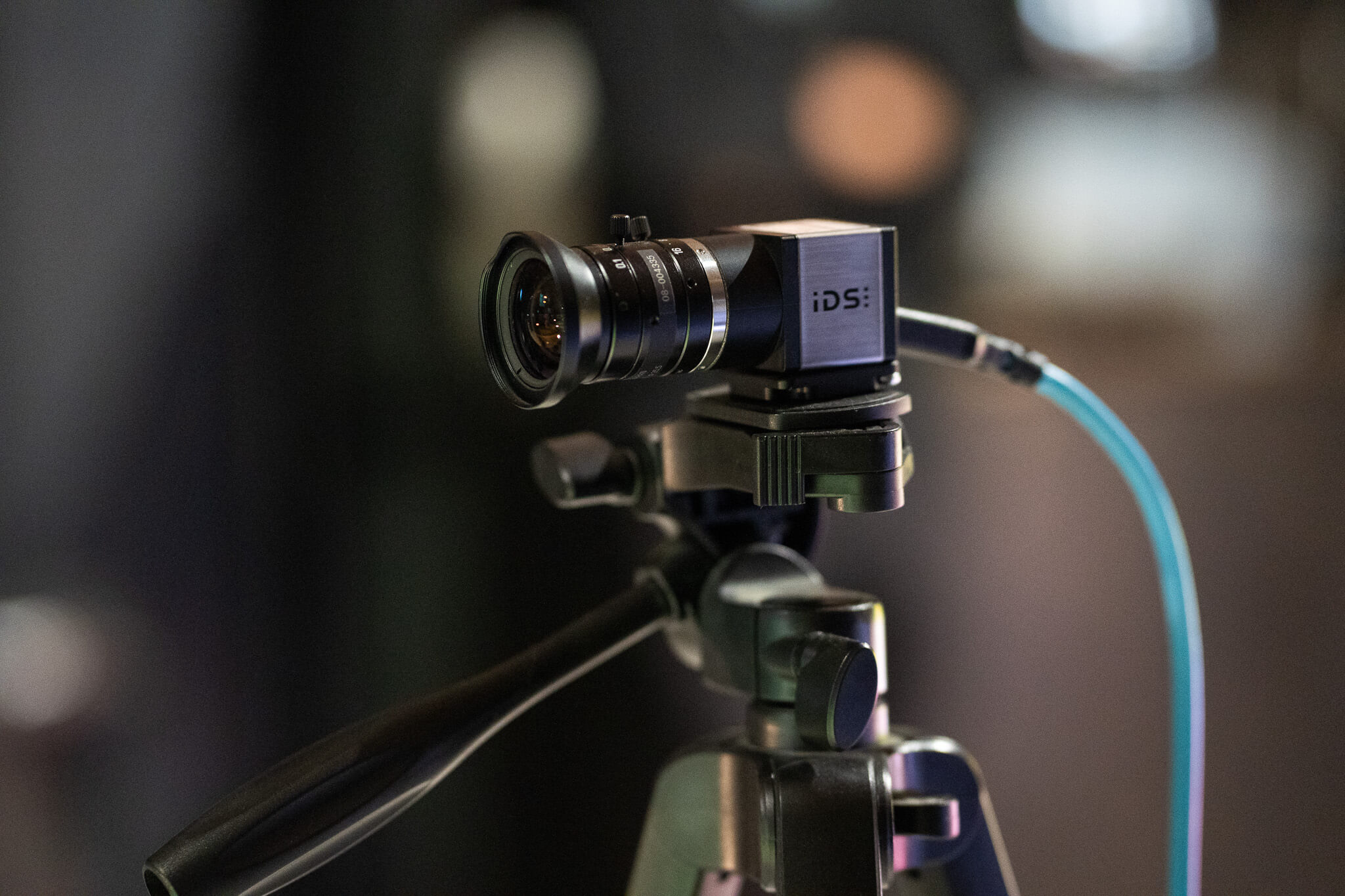 A close-up of a compact iDS camera mounted on a tripod, with a blue cable connected to the camera, set against a blurred background.