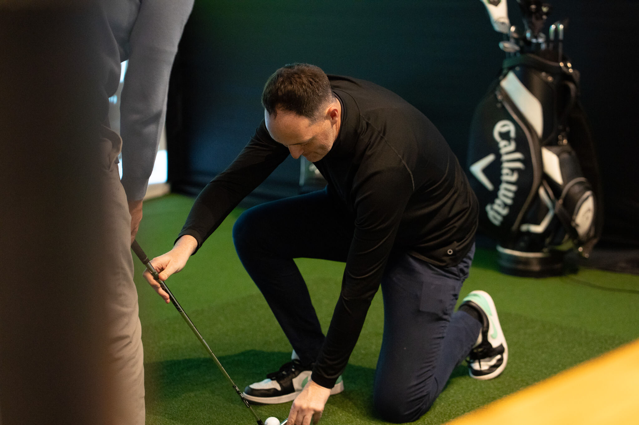 A man in black sportswear kneels on artificial turf, positioning a golf ball and putter during golf lessons. A golf bag is visible in the background, while another person stands nearby, partially out of frame.