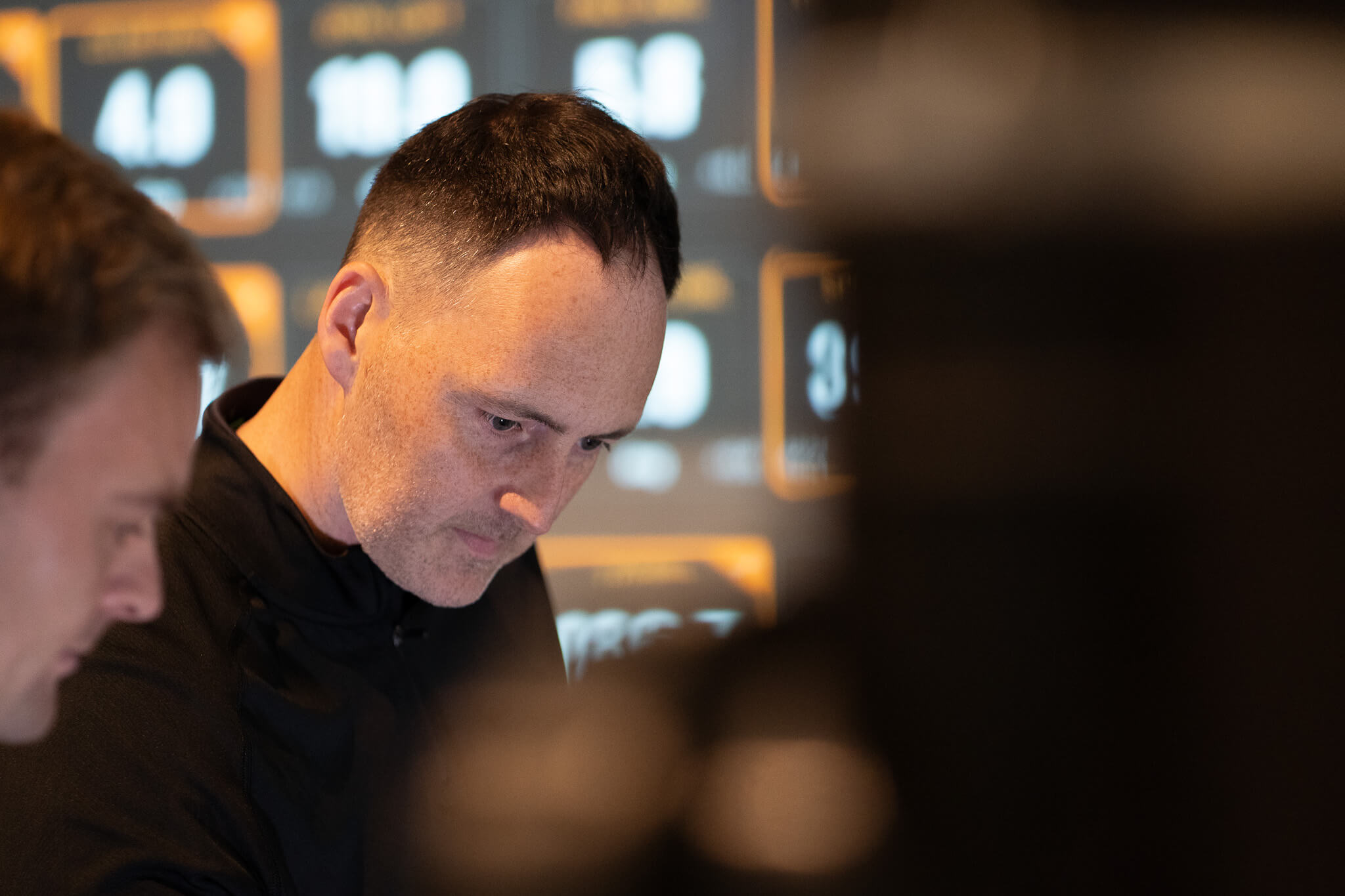 A man with short dark hair looks down with a serious expression. Blurred TrackMan screens displaying numbers and golf data are visible in the background, and another person is partially visible on the left.