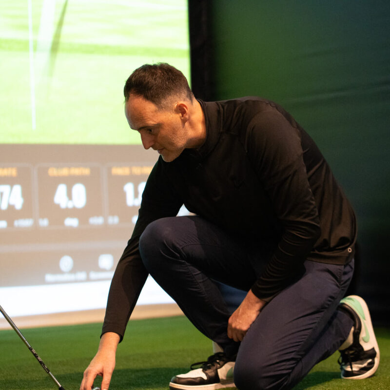 A man kneels on artificial turf indoors, adjusting a golf ball near a golf club. A large screen displays TrackMan golf statistics in the background, and a golf bag stands nearby, capturing the immersive experience of indoor golf London.