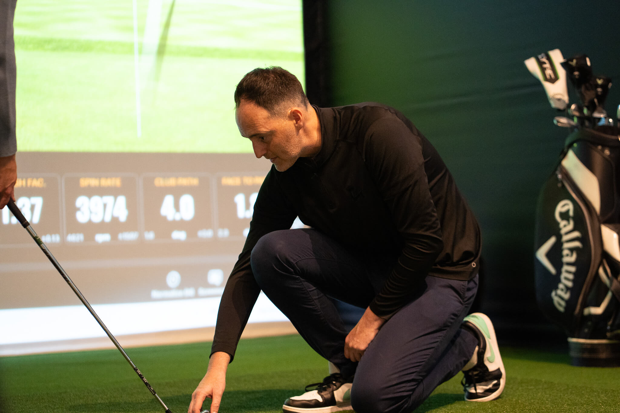 A man kneels on artificial turf indoors, adjusting a golf ball near a golf club. A large screen displays TrackMan golf statistics in the background, and a golf bag stands nearby, capturing the immersive experience of indoor golf London.