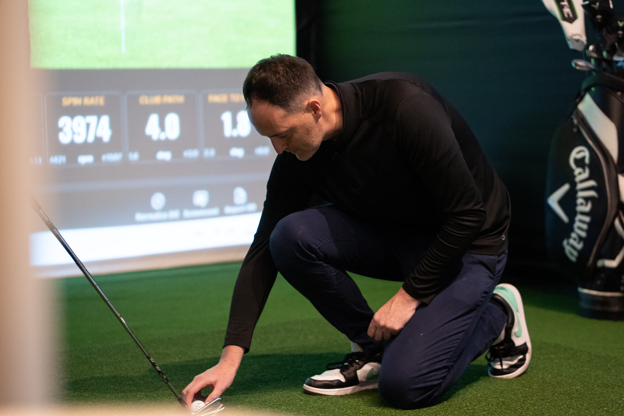 A man in a black long-sleeve shirt and blue pants kneels on artificial turf, adjusting a golf ball near a club. A golf bag and digital golf simulator screen highlight this indoor golf London experience.