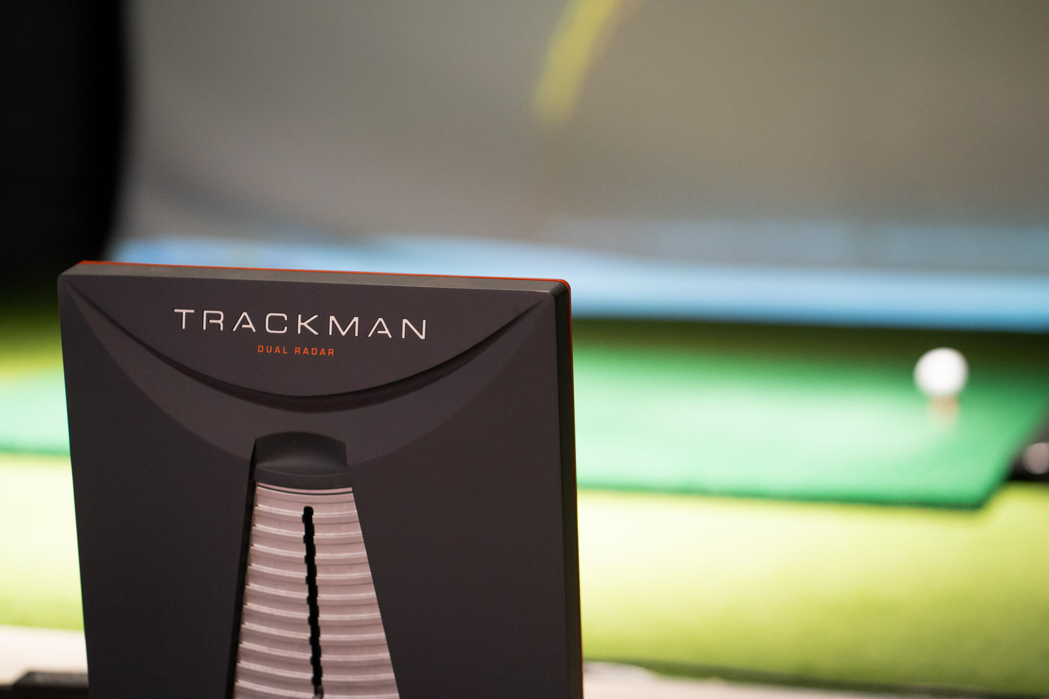 A TrackMan radar device is set up indoors, focused on a golf ball positioned on a mat.