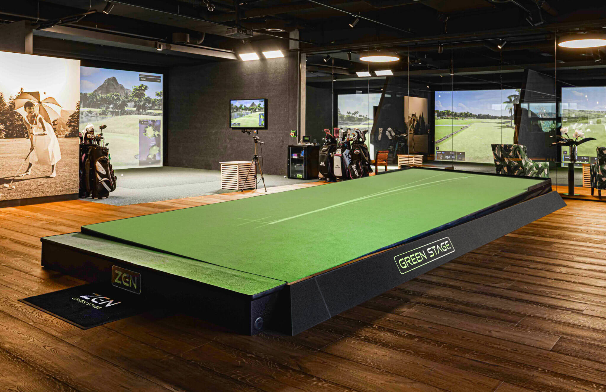 A modern indoor golf simulation room in London featuring a raised green putting stage, golf bags, and large screens displaying golf courses. The space has wooden flooring, golf-themed wall art, and options for golf lessons or membership.