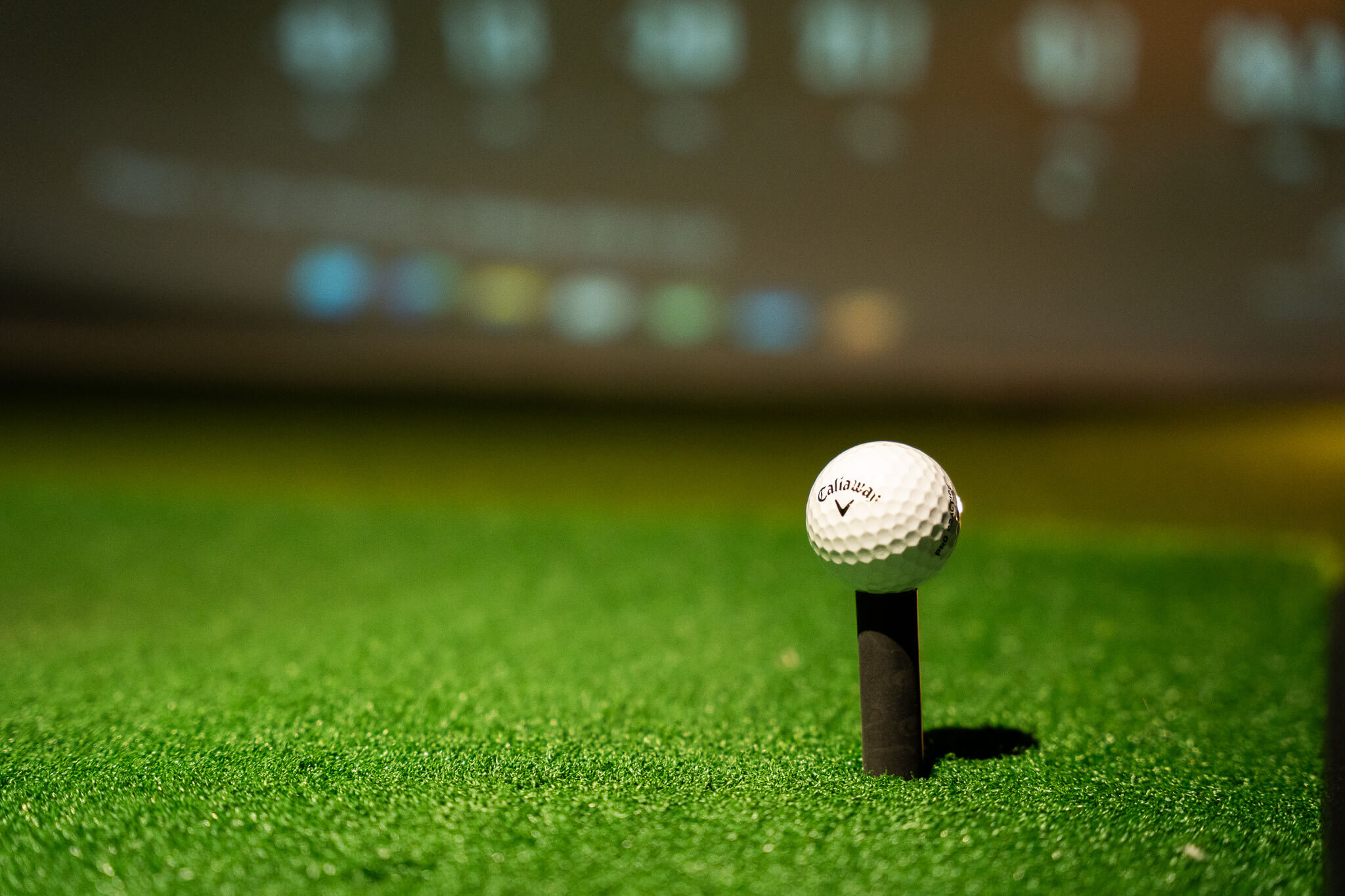A close-up of a Callaway golf ball placed on a black tee, set on bright green artificial turf at a golf lesson, with a blurred screen displaying colorful lights and numbers in the background.