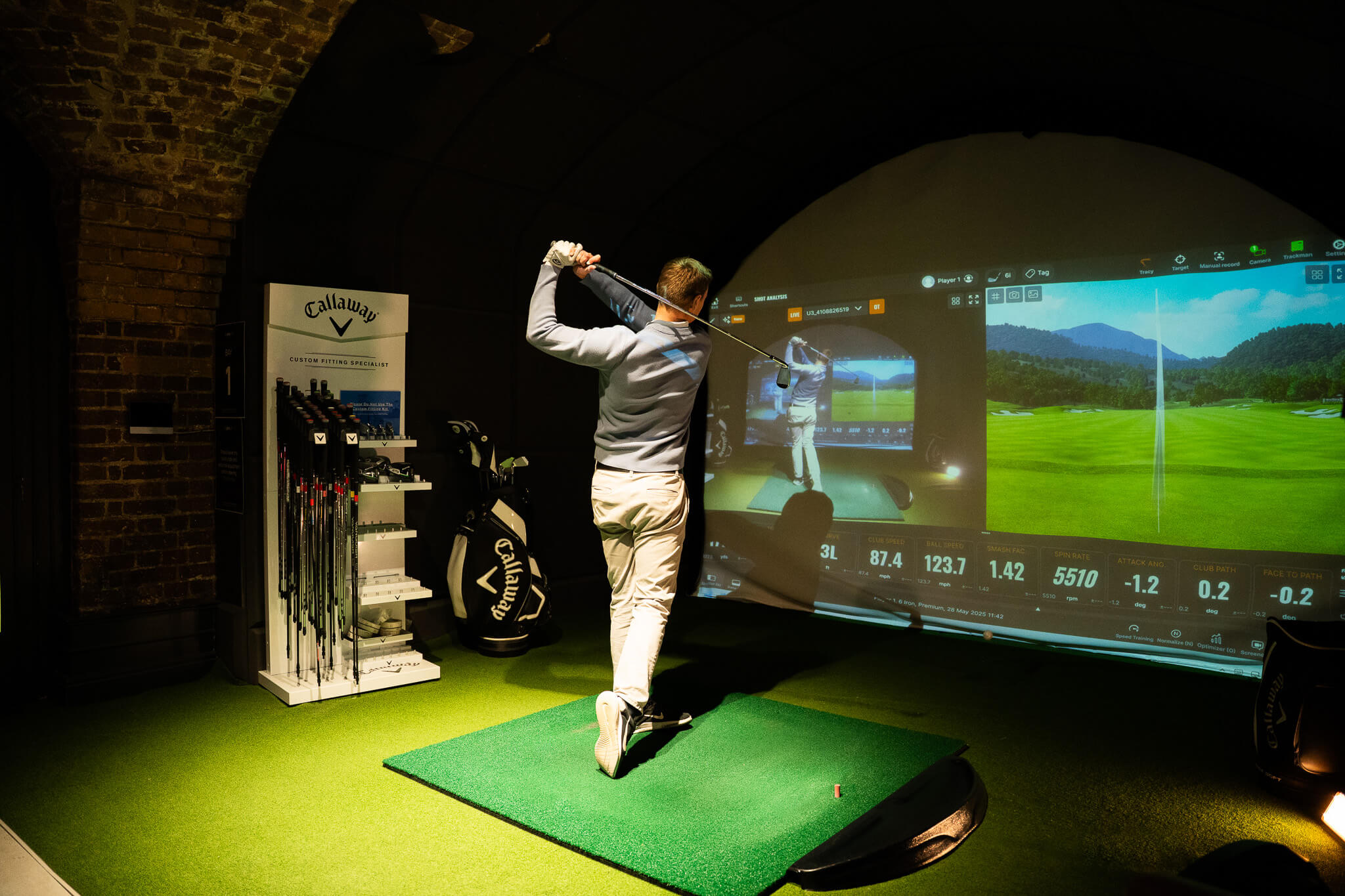 A person swings a golf club in an indoor golf simulator, facing a large screen displaying a virtual golf course and swing data—perfect for golf lessons or practicing your game. Golf clubs and bags are visible on the left.