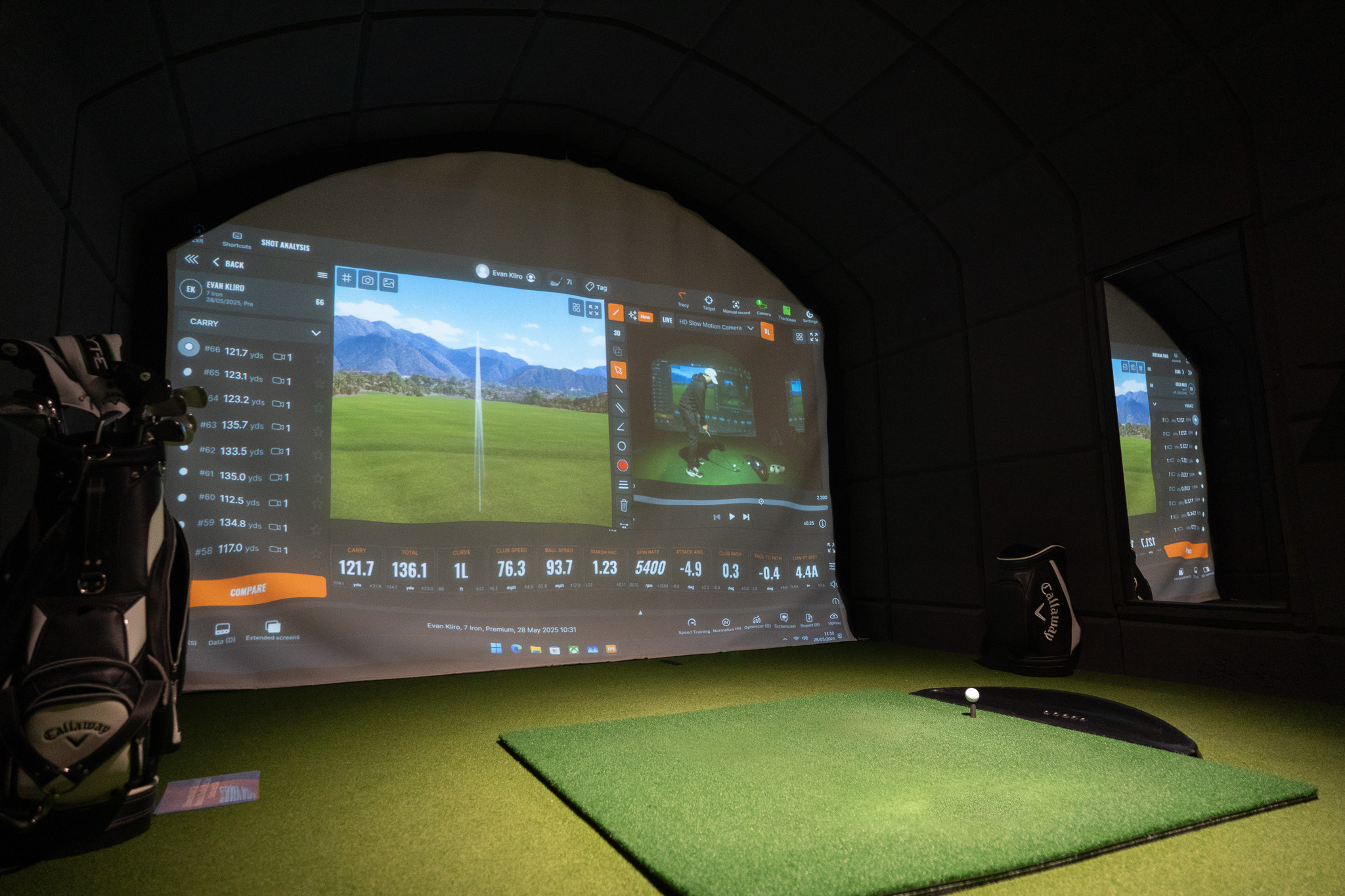 A golf simulator room featuring TrackMan technology, a large screen displaying golf swing data and a virtual golf course, artificial turf flooring, a golf ball on a mat, a golf bag, and a mirror reflecting the scene—perfect for any golf membership.
